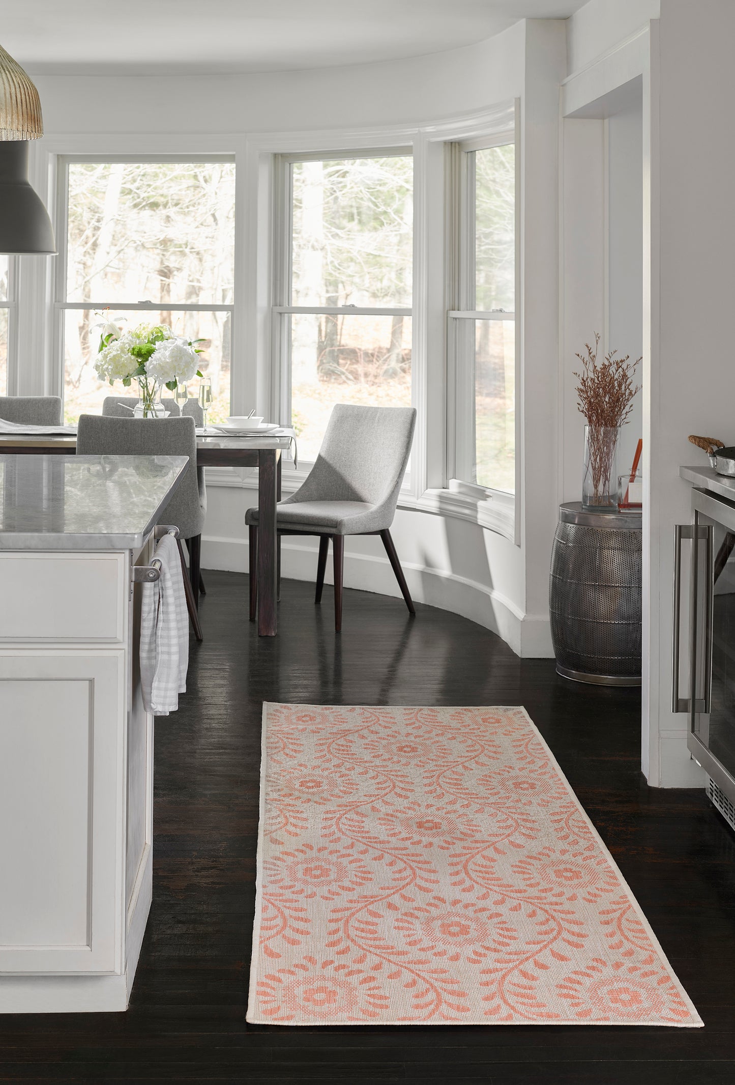 Bright open kitchen with white cabinets, dark wood floors, marble island, curved bay windows, peach-patterned rug.