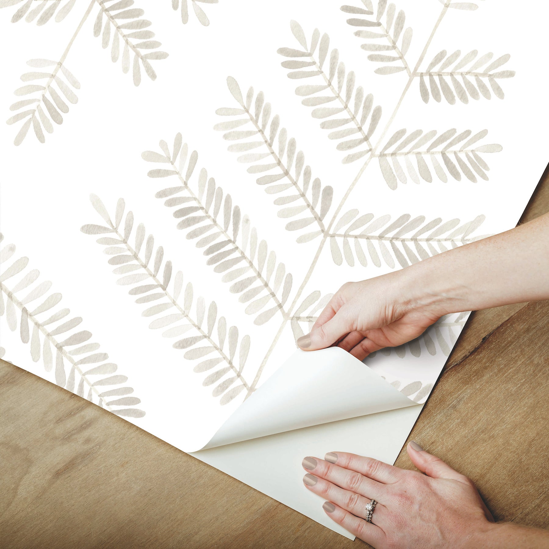 Beige fern leaf wallpaper sample being peeled back to reveal a curled corner on a wooden surface.