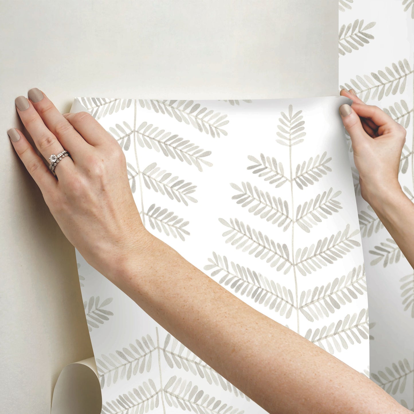 Hands peel a white wallpaper sheet with a pale gray leaf pattern, modern botanical wall decor.
