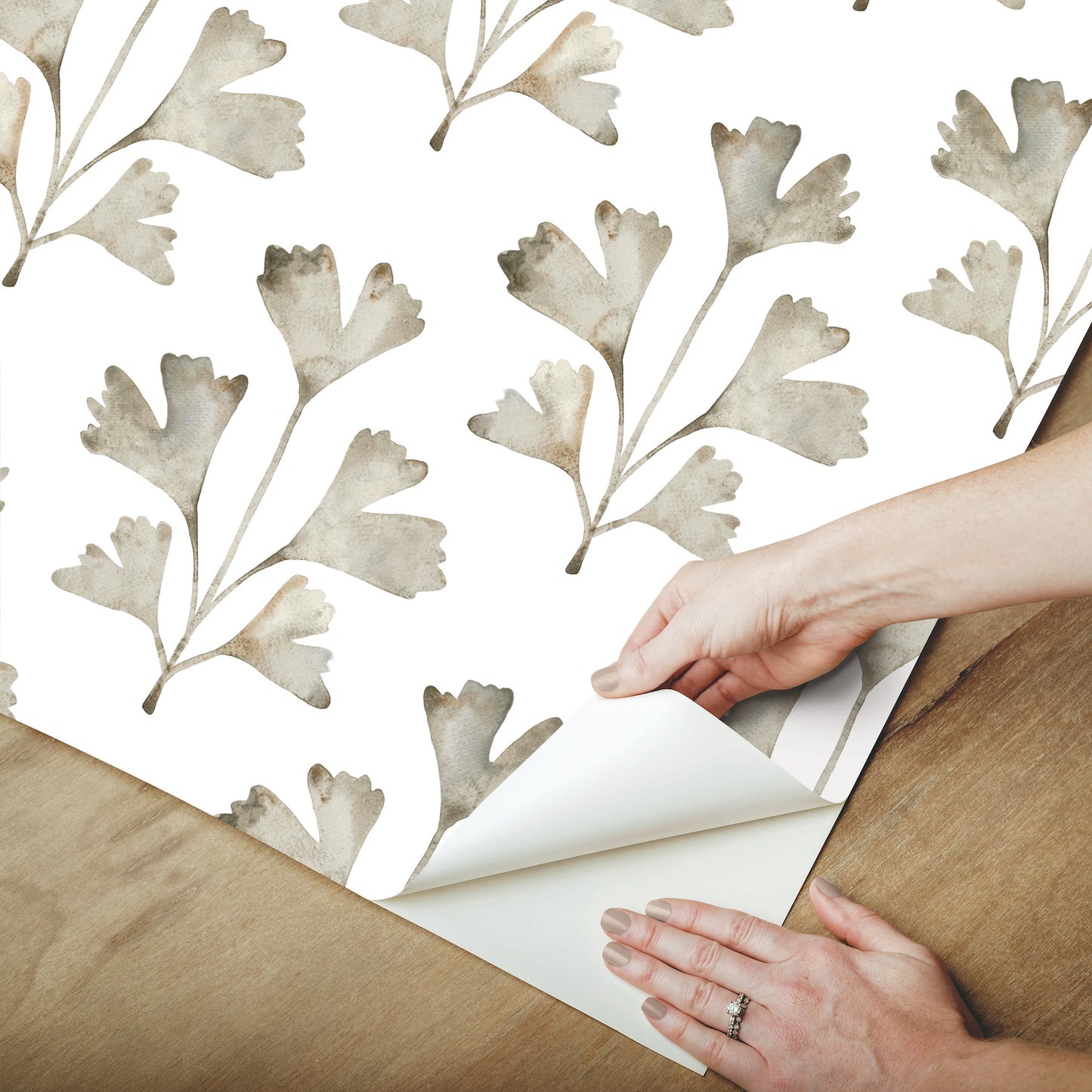 Hands peeling back neutral gray botanical leaf wallpaper on white background, watercolor leaves, wooden floor visible.