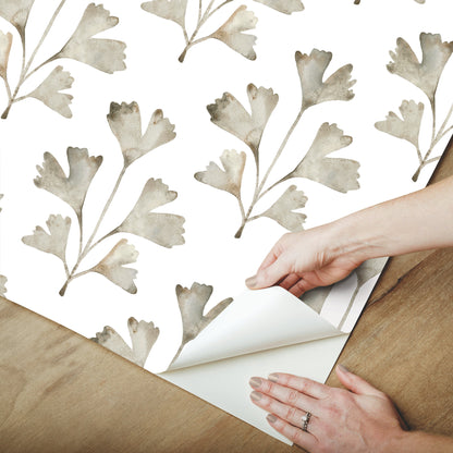 Hands peeling back neutral gray botanical leaf wallpaper on white background, watercolor leaves, wooden floor visible.