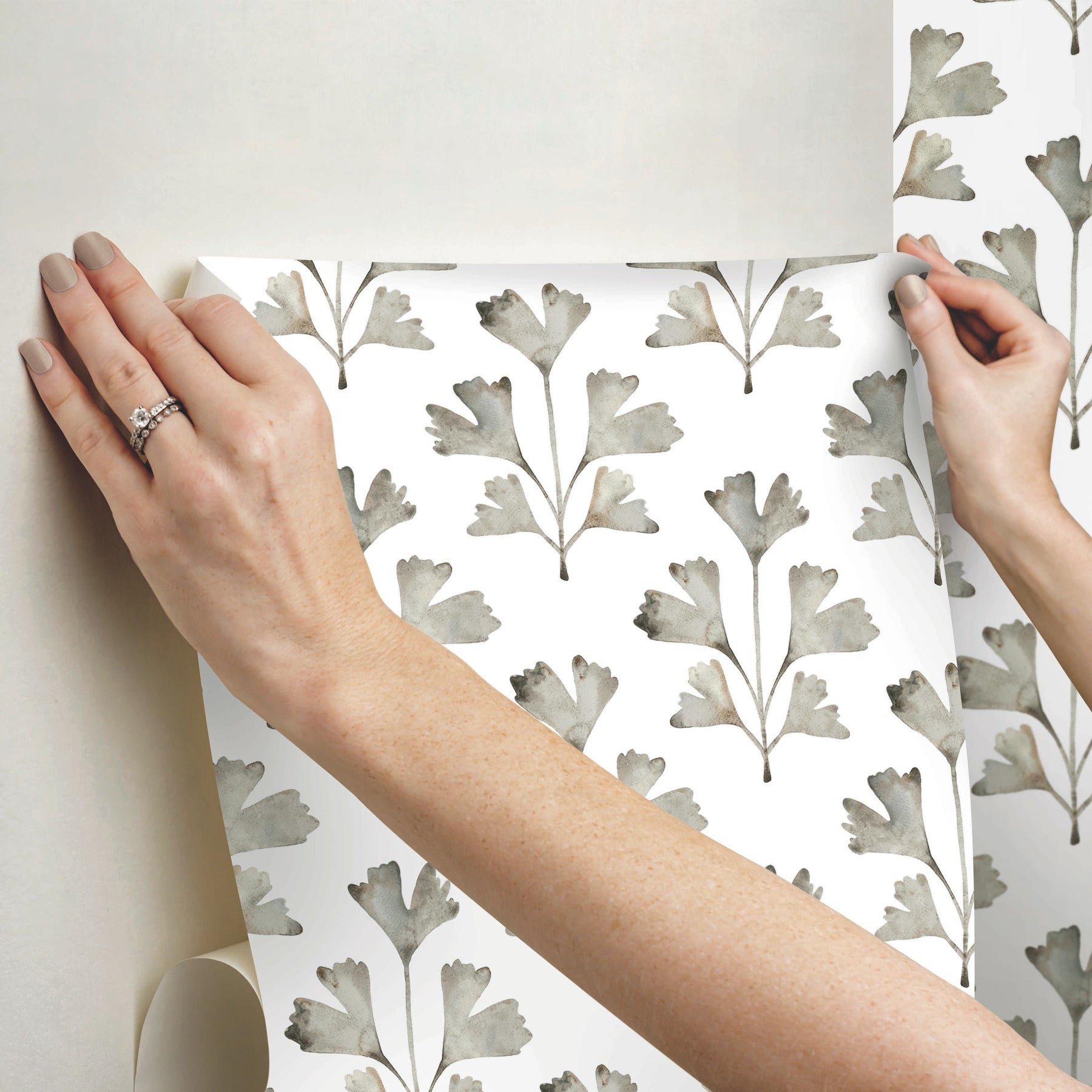 Hands hold a sheet of white wallpaper featuring a muted gray botanical leaf pattern.
