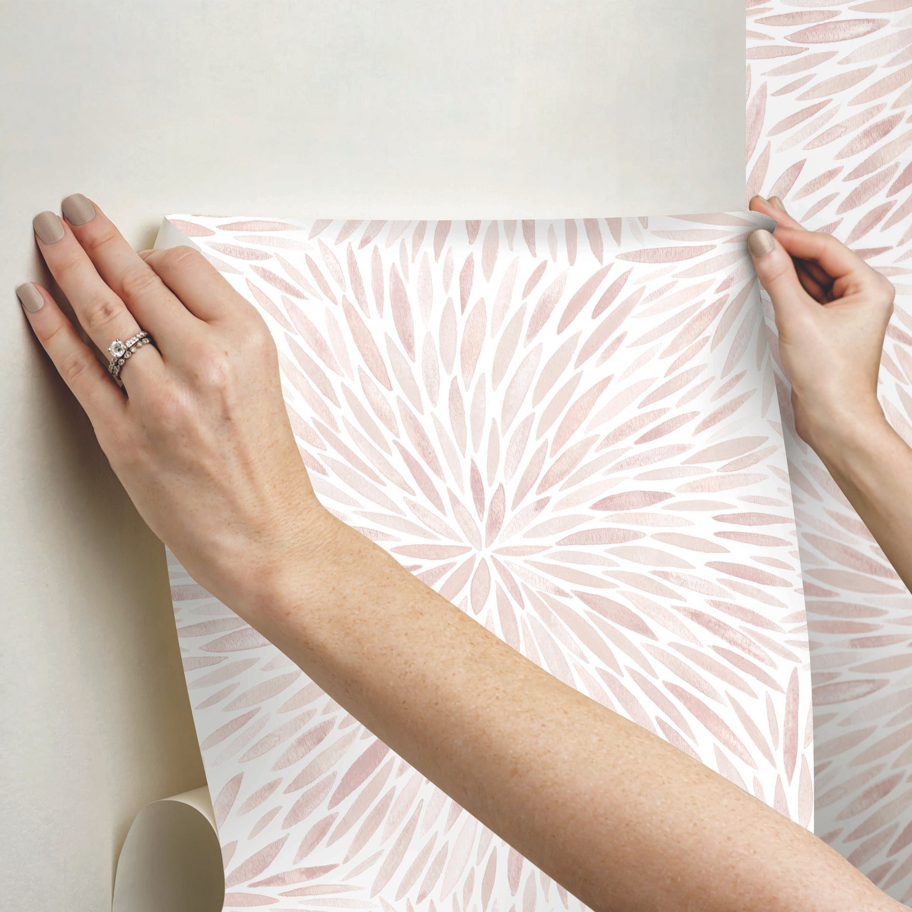 Hands hold a soft pink botanical wallpaper sample with a radiating leaf motif against a pale background.