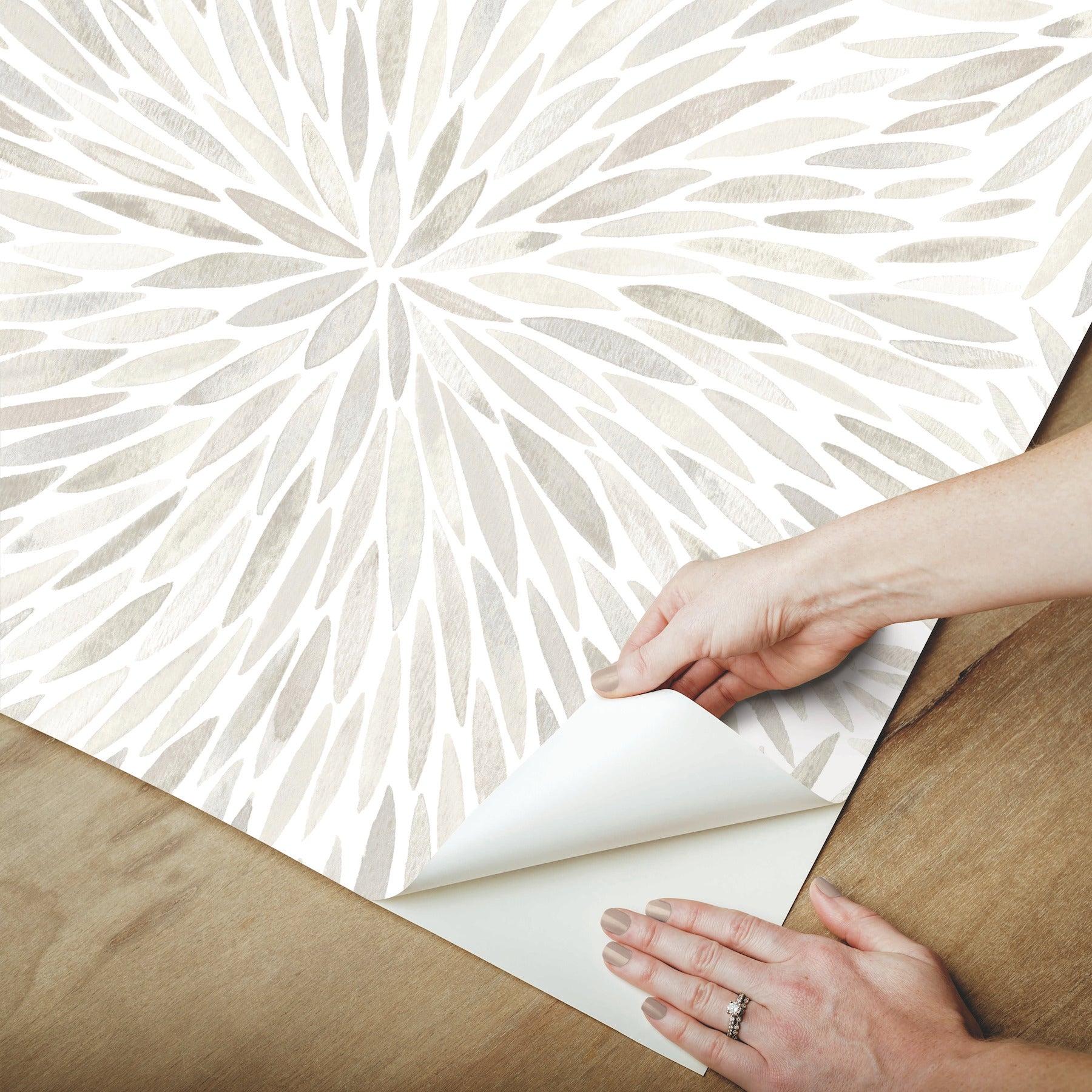 Hands peel back corner of light gray abstract leaf wallpaper on a wooden floor.