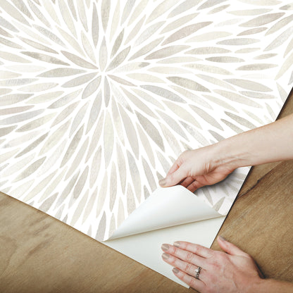 Hands peel back corner of light gray abstract leaf wallpaper on a wooden floor.