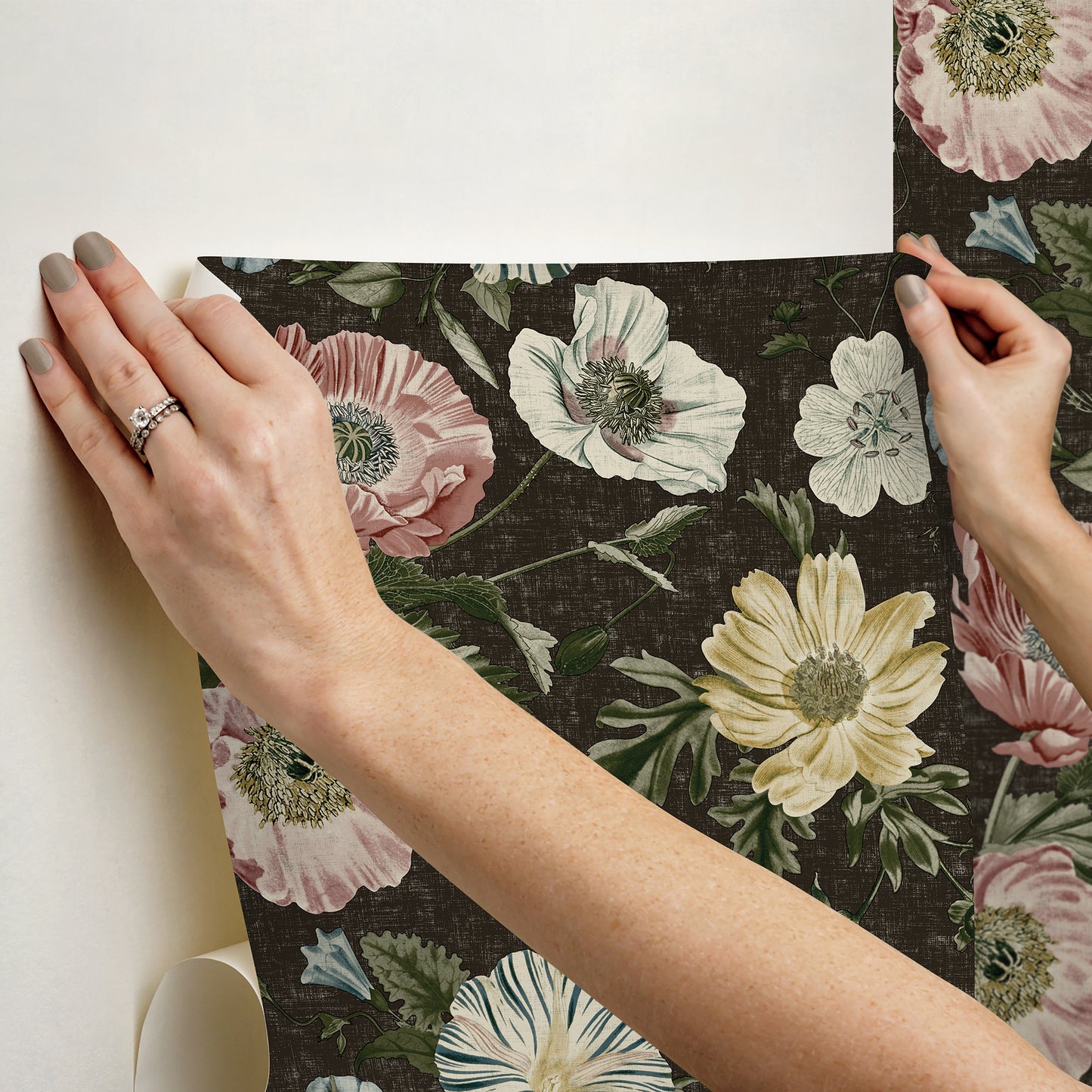 Two hands hold a dark floral wallpaper panel showing large pale pink, white, and yellow flowers; vintage botanical design.