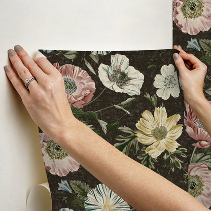 Two hands hold a dark floral wallpaper panel showing large pale pink, white, and yellow flowers; vintage botanical design.