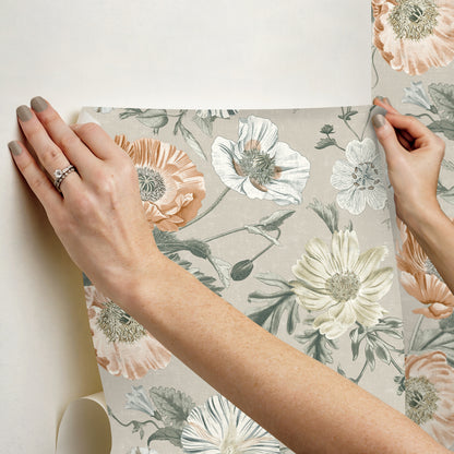 Two hands hold a floral wallpaper sample in peach, cream, and sage on a light gray background.