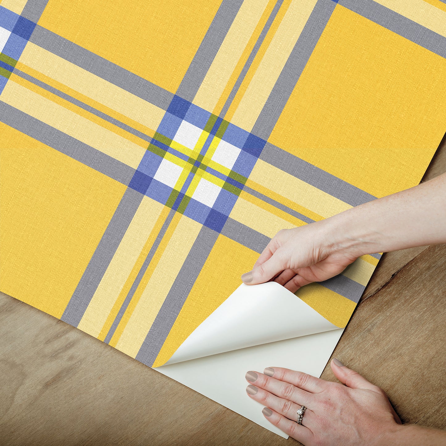 Yellow plaid wallpaper sample with blue and gray lines being peeled back at corner on wood surface.