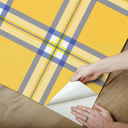 Yellow plaid wallpaper sample with blue and gray lines being peeled back at corner on wood surface.