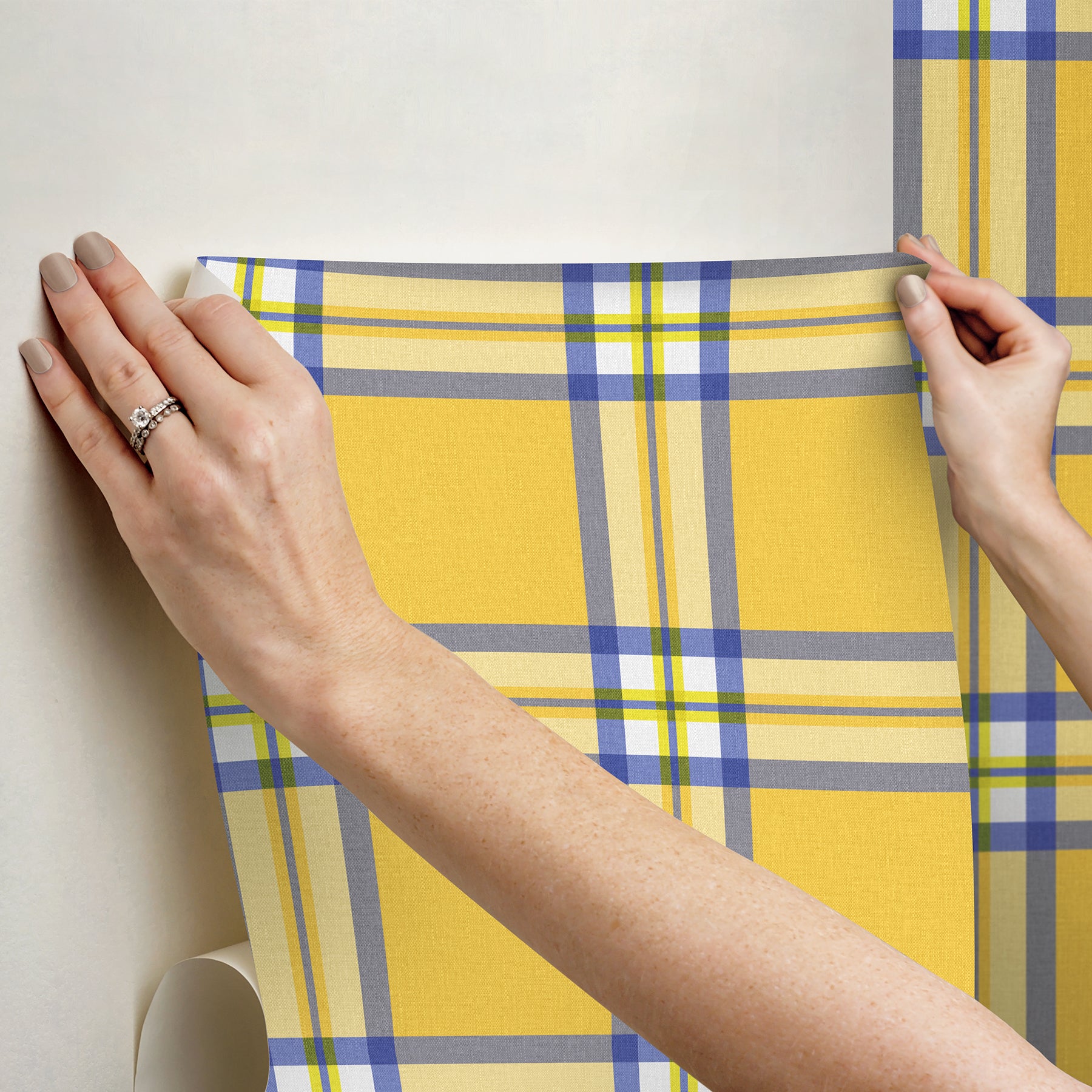 Yellow plaid wallpaper sheet with blue and gray stripes, held and unrolled by two hands.
