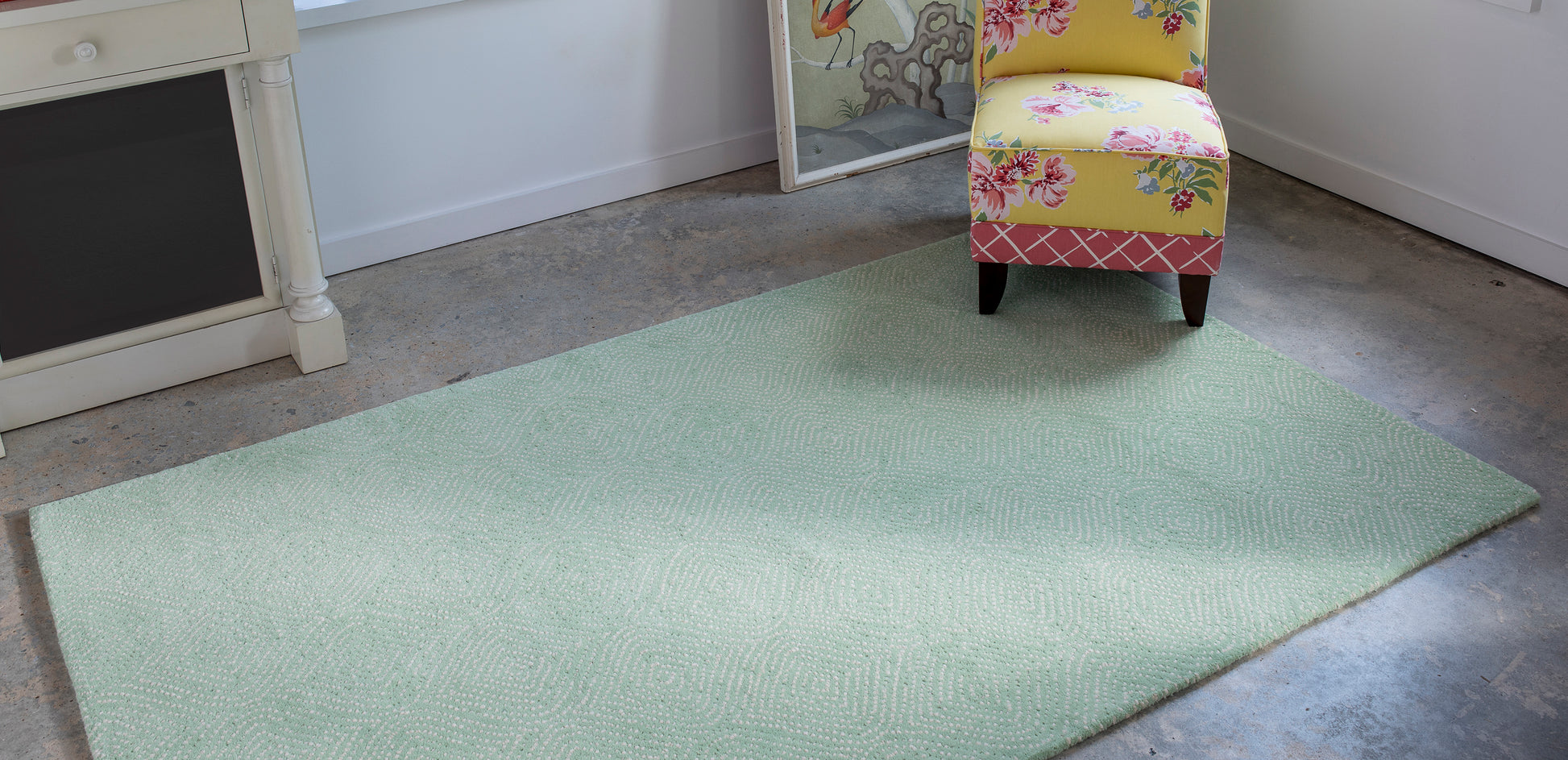 Mint green rectangular flatwoven rug on concrete floor in a bright room with a floral upholstered chair.