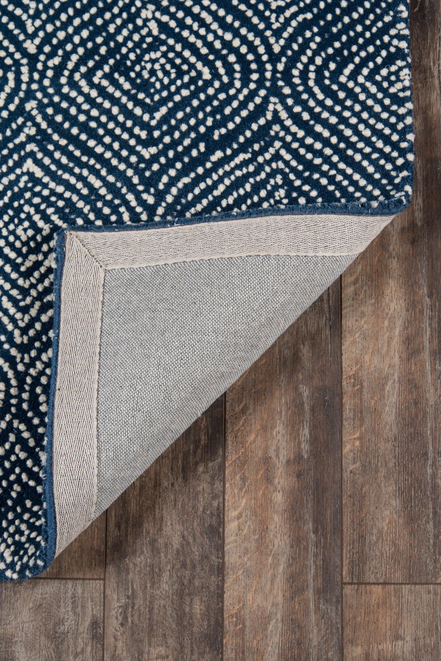 Navy woven rug with white dotted pattern; corner folded to show light gray backing on wood floor.
