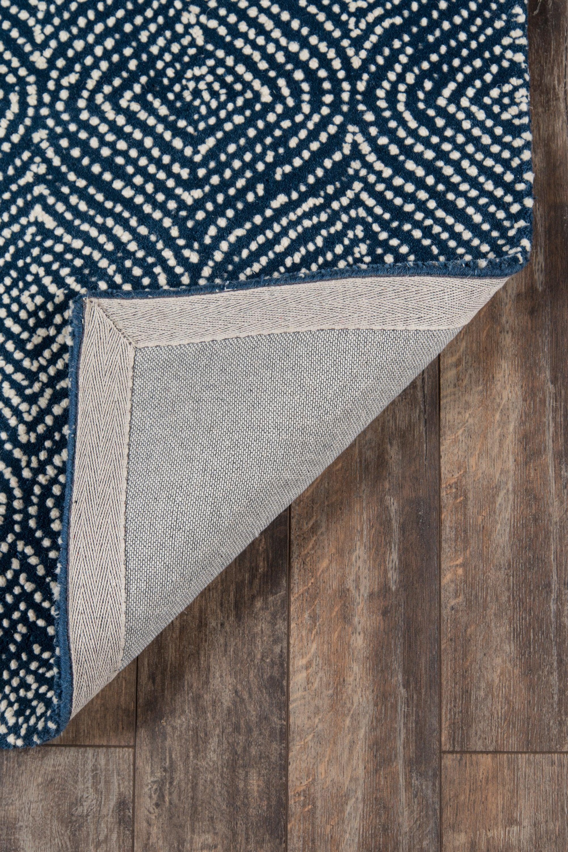 Navy woven rug with white dotted pattern; corner folded to show light gray backing on wood floor.