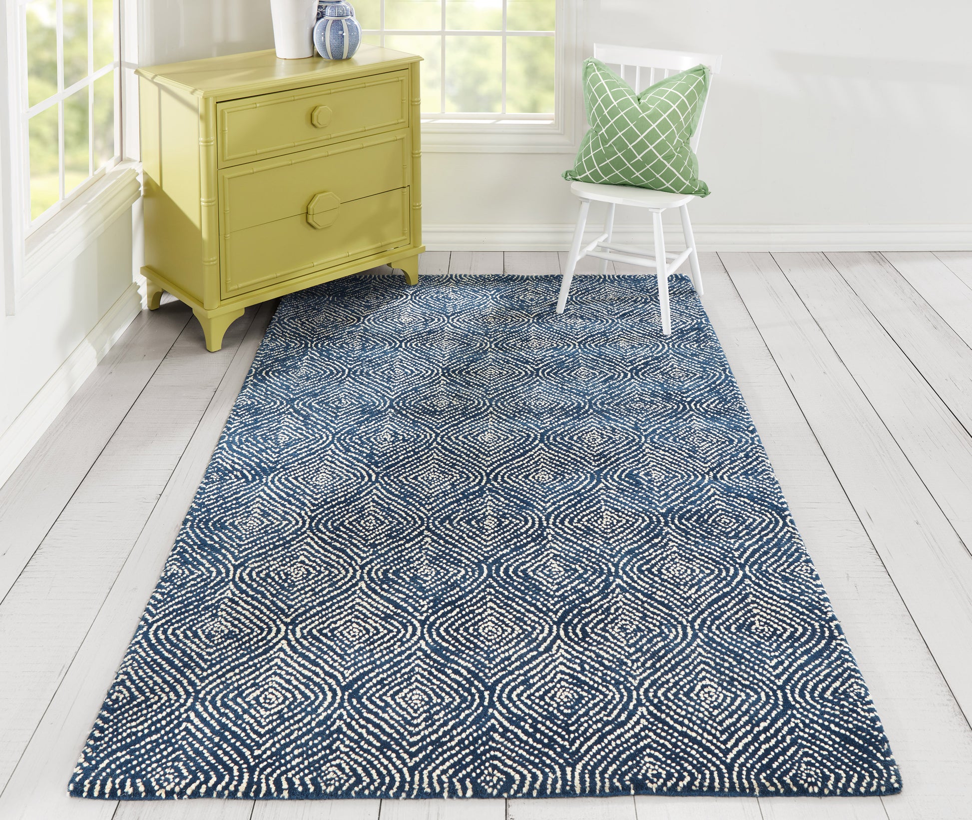 Navy blue rug with a white geometric pattern on a light wood floor beside a yellow dresser & a chair with a green pillow.