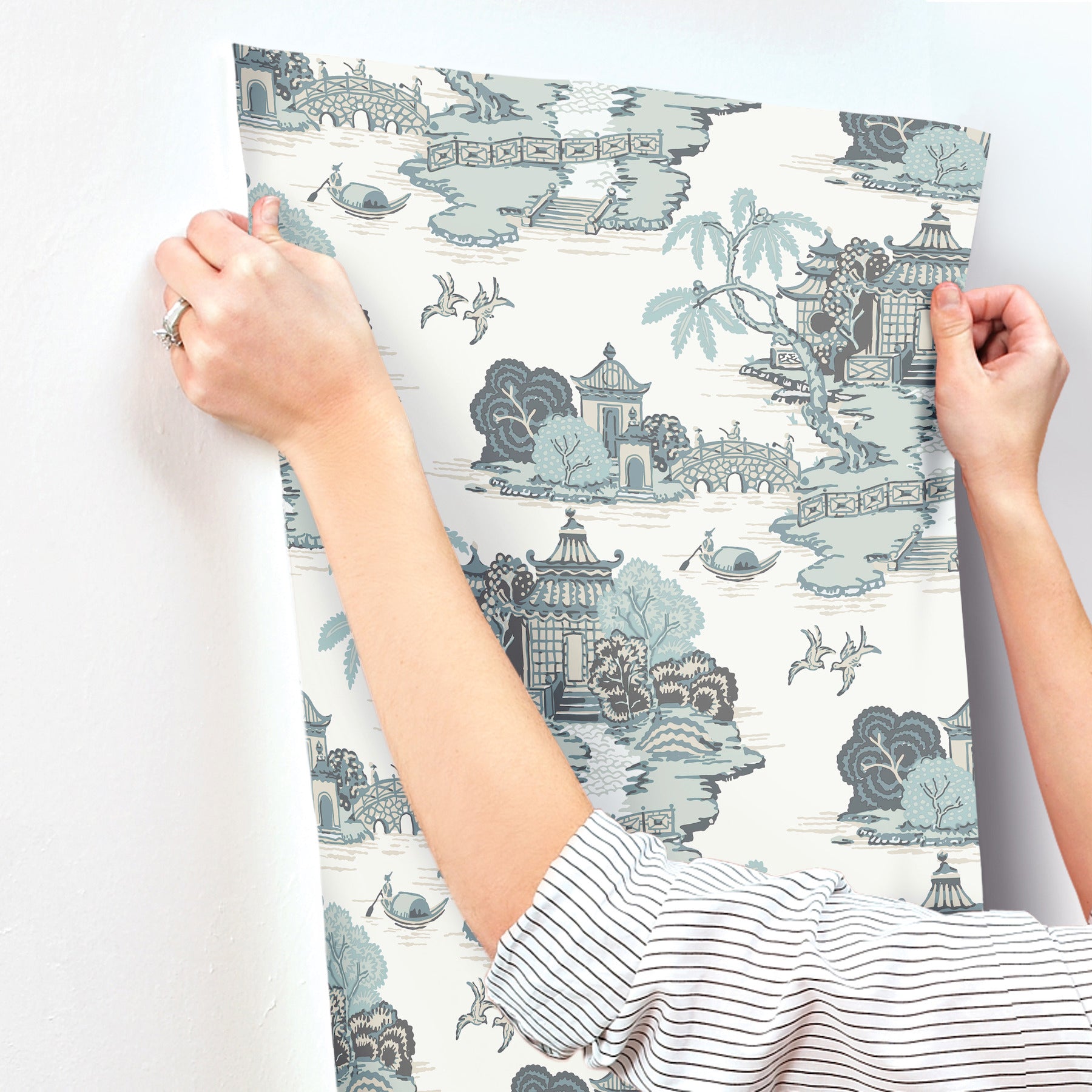 Person holding blue-gray toile wallpaper panel featuring pagodas, bridges, boats and trees.