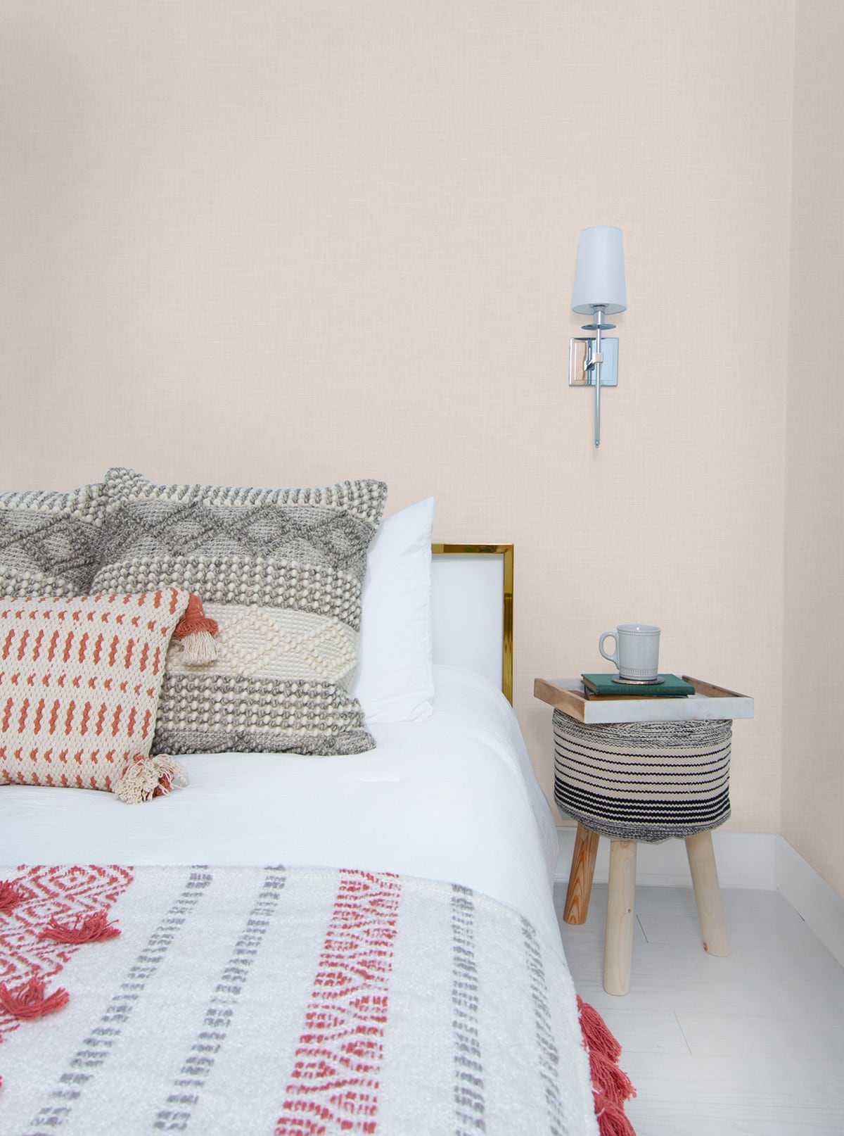 Cozy bedroom with white bedding, patterned throw pillows, striped fabric side table, coffee mug and book, wall sconce; beige matte embossed vinyl wallpaper on the walls.