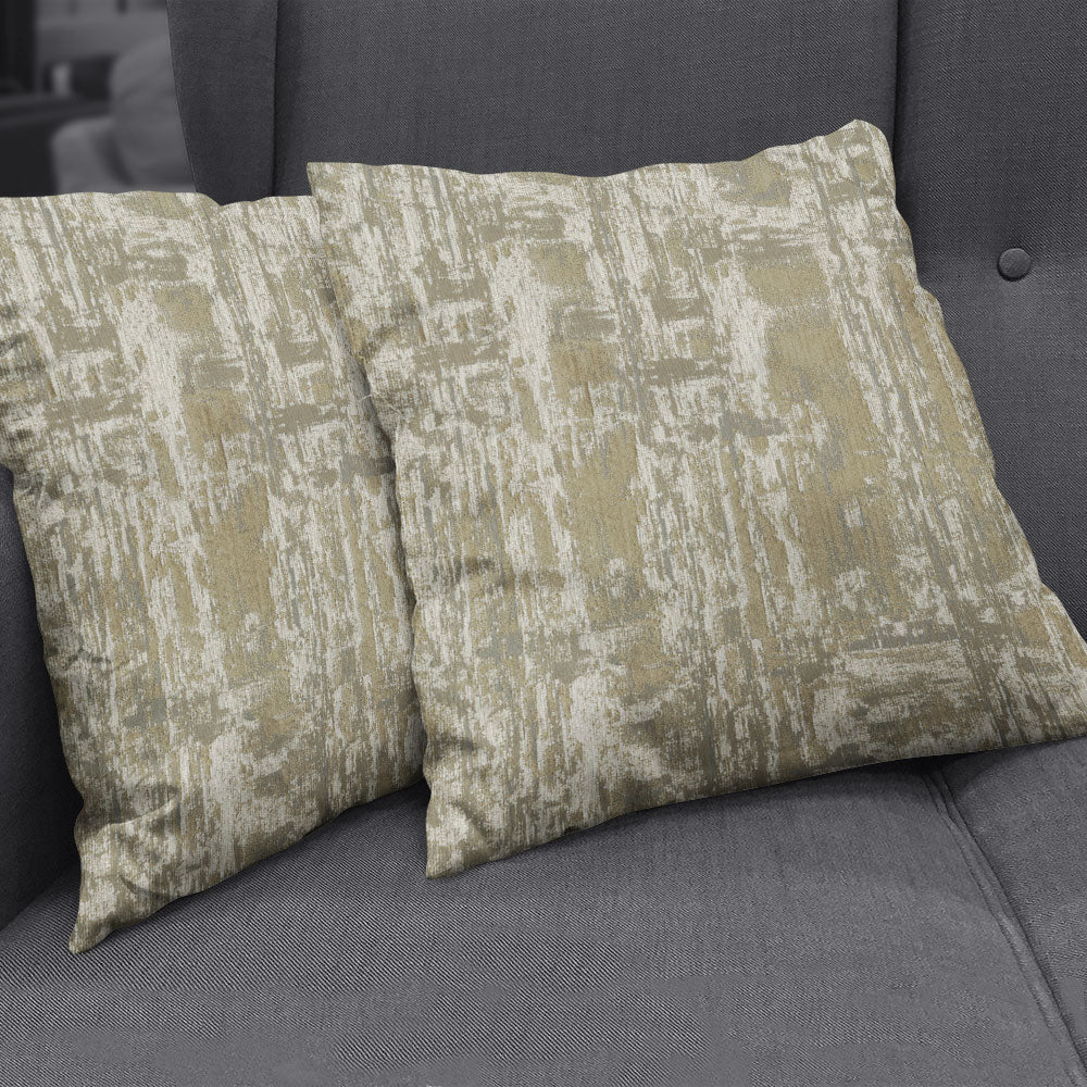 Two square decorative throw pillows with a beige and pale gold hemp abstract pattern on soft chenille/velvet fabric on a gray sofa