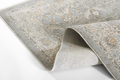 Blue-gray rug with ornate pattern and subtle gold accents, folded corner reveals light backing.