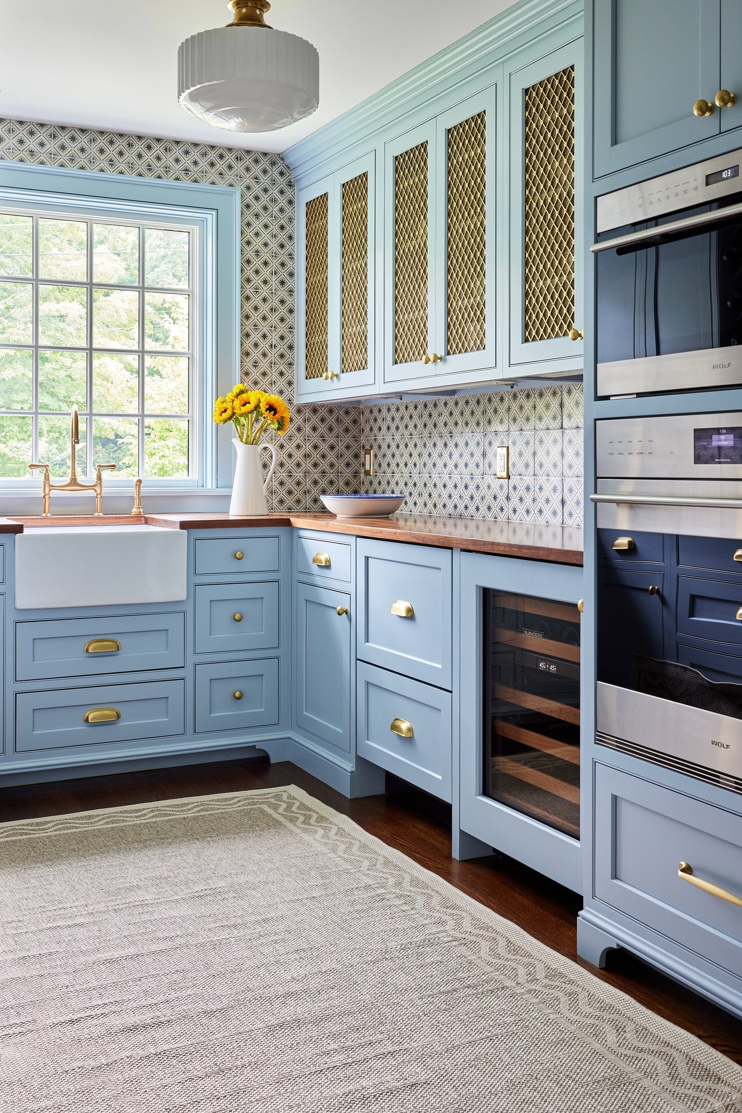 Blue-gray kitchen with brass hardware, glass-front lattice doors, built-in ovens, wine fridge, and yellow flowers.