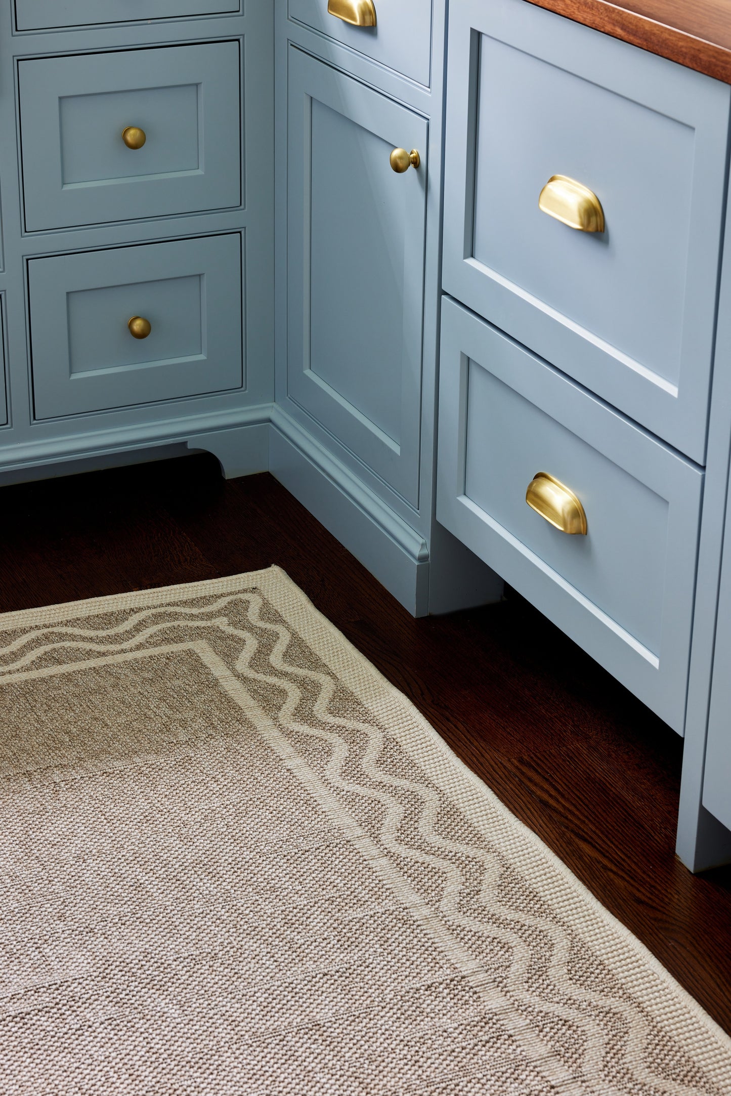 Blue-gray shaker cabinets with brass cup pulls, wood countertop, corner base cabinets, dark hardwood and beige patterned rug.