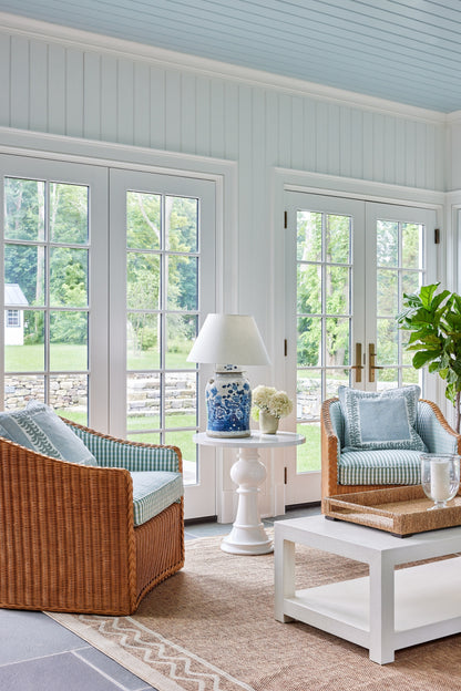 Bright coastal living room with blue/white striped cushions on wicker chairs, white coffee table, ceramic lamp, jute rug.