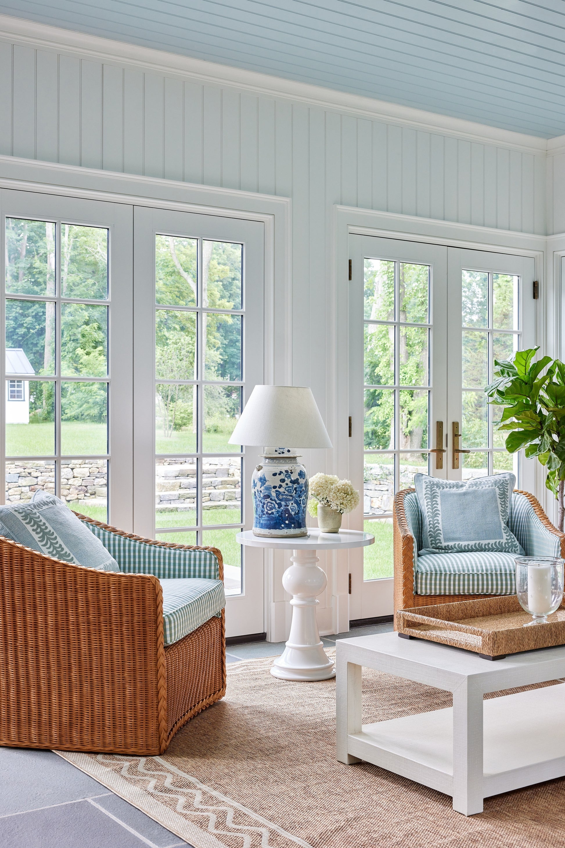 Bright sunlit living room with wicker chairs, blue cushions, white coffee table, blue-and-white lamp, plants, glass doors.