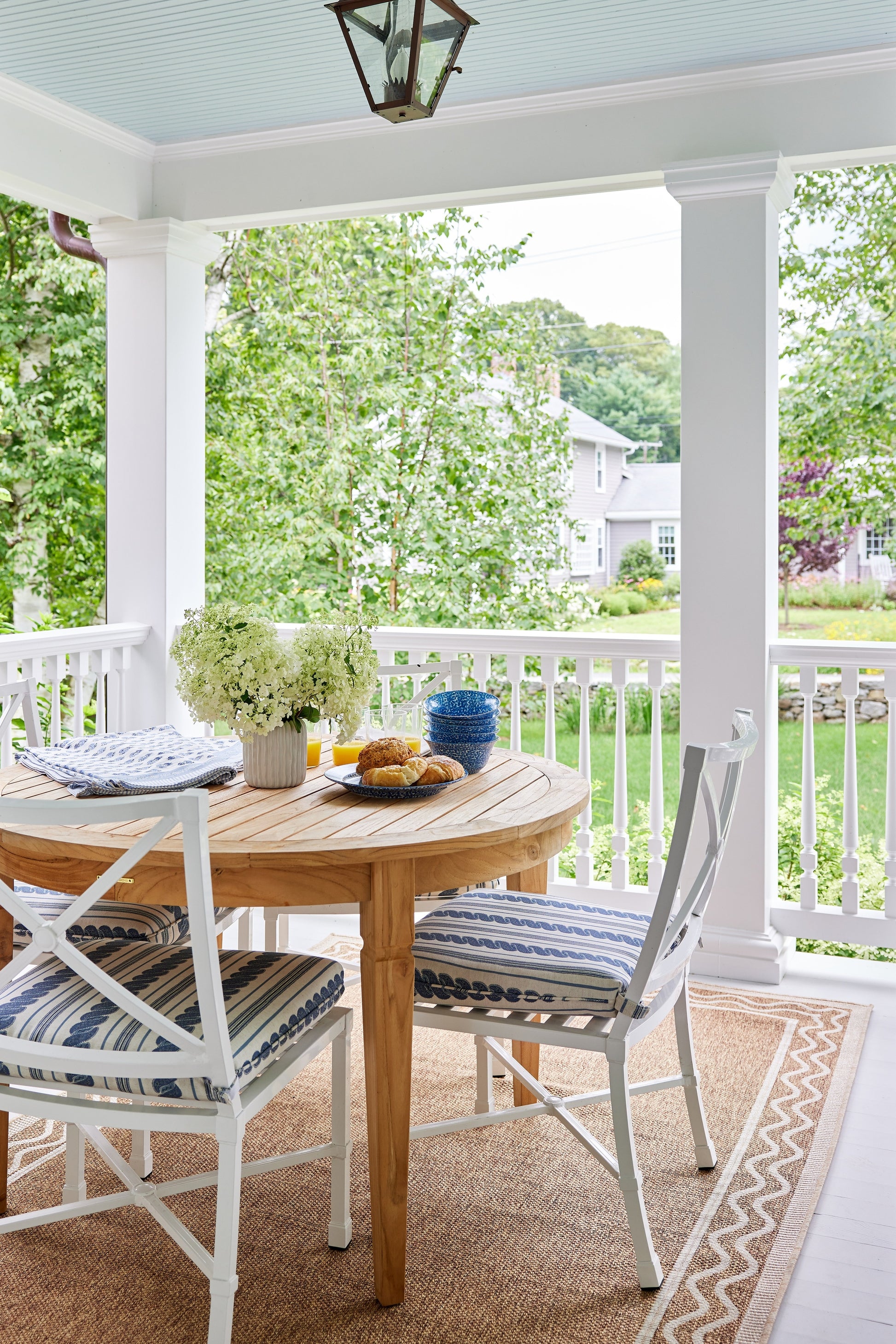 Outdoor porch dining set: round natural-wood table, white chairs with blue striped cushions, hydrangea centerpiece.