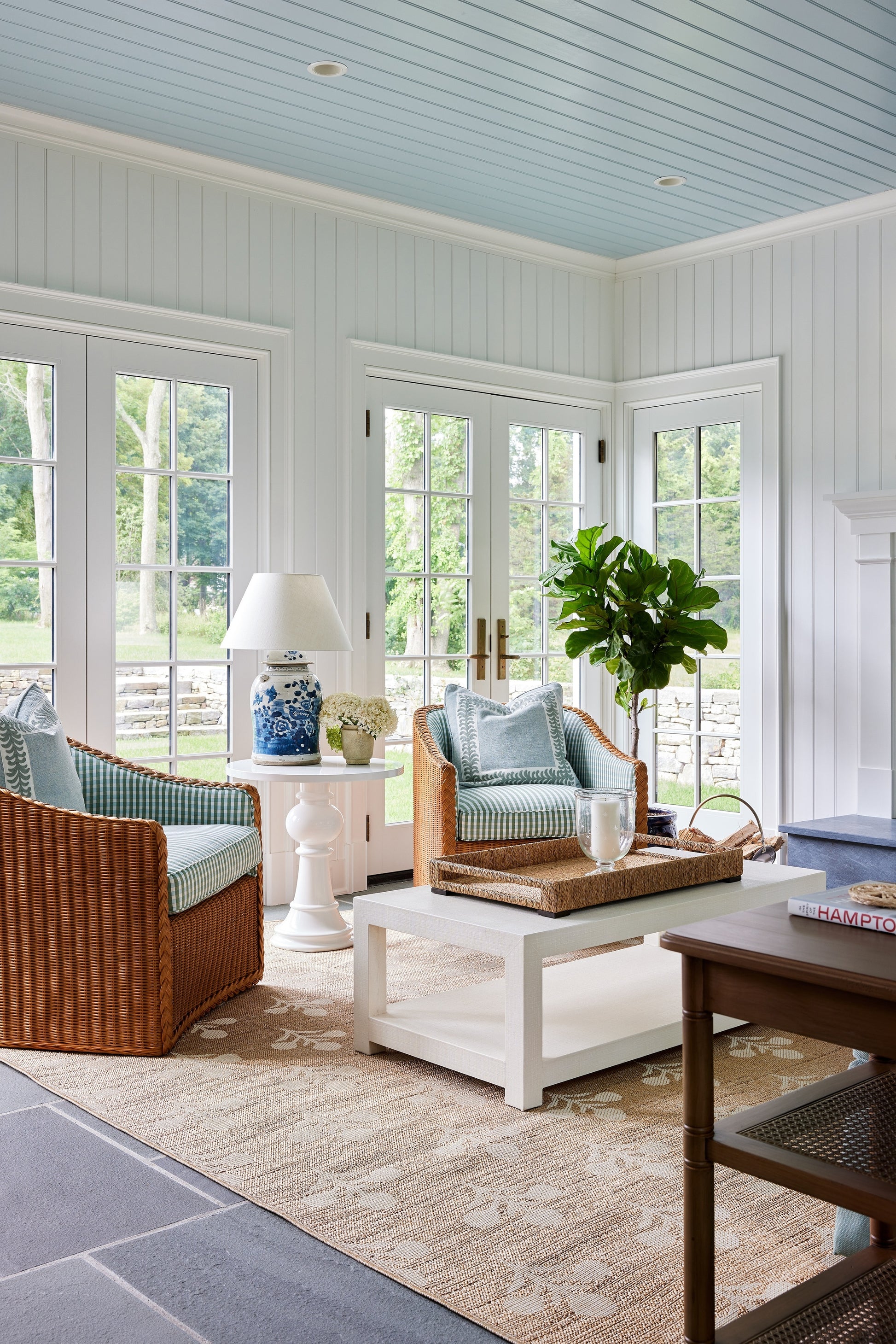 Bright coastal sunroom featuring a rug, wicker armchairs with blue-green cushions, a white coffee table, a potted plant, a lamp and glass doors.