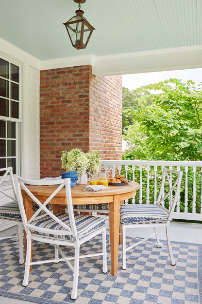 Round wooden outdoor dining table with white wicker chairs and blue cushions, hydrangea bouquet on blue-check porch tiles.