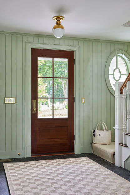 Dark wood door with glass panes, sage beadboard walls, arched window, brass light, checkered rug, white stairs, tote bag