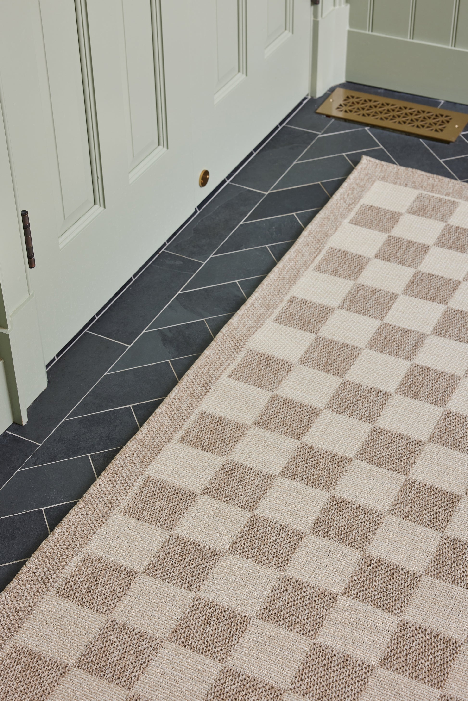 Beige and taupe checkerboard runner rug on dark gray chevron tile floor by a pale-green door.