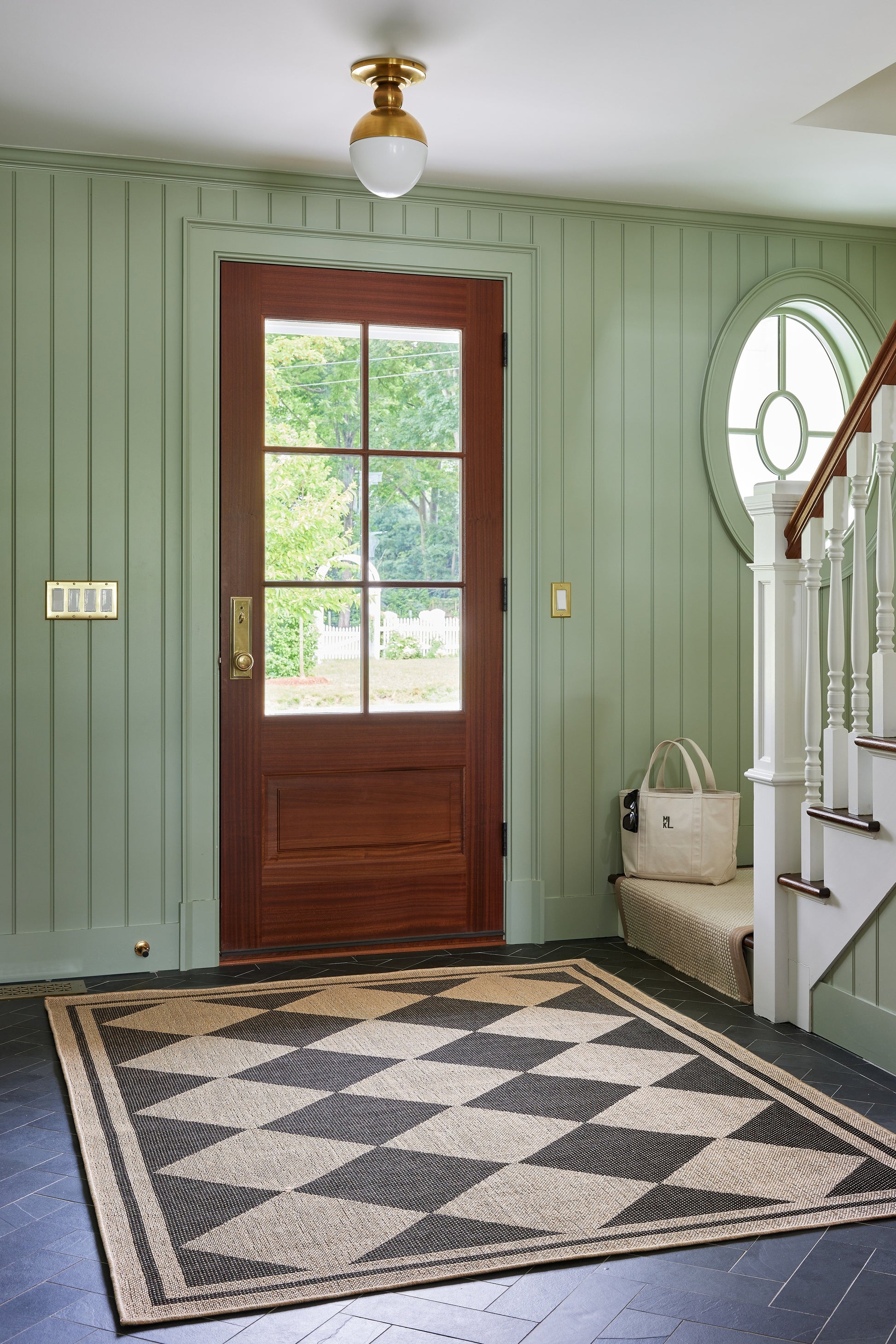 Mint green beadboard entryway with dark wood door, black-and-cream geometric rug, cream bench, beige bag, circular window by stairs.