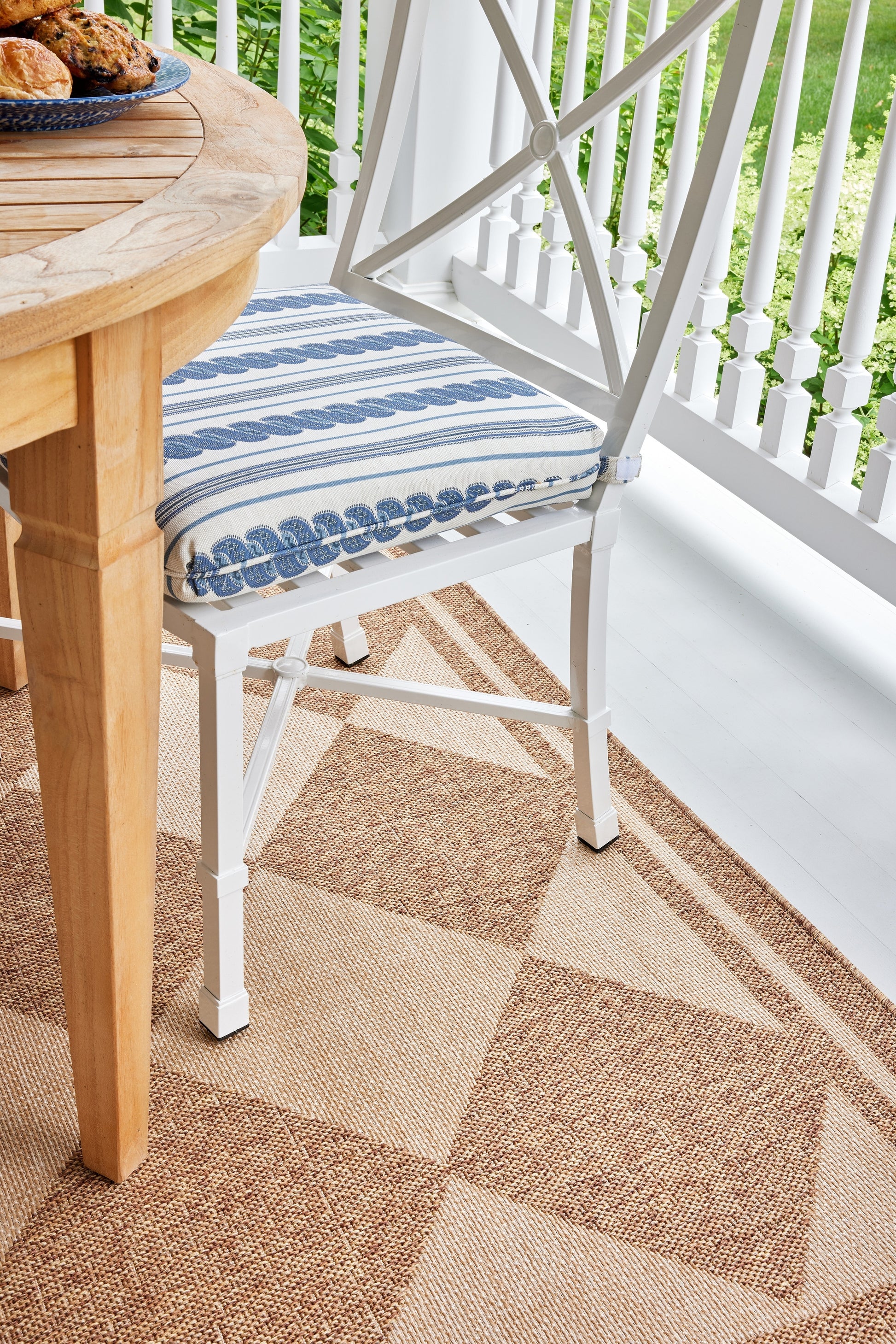 White metal outdoor chair with blue striped cushion beside a wooden round table on a beige geometric outdoor rug.