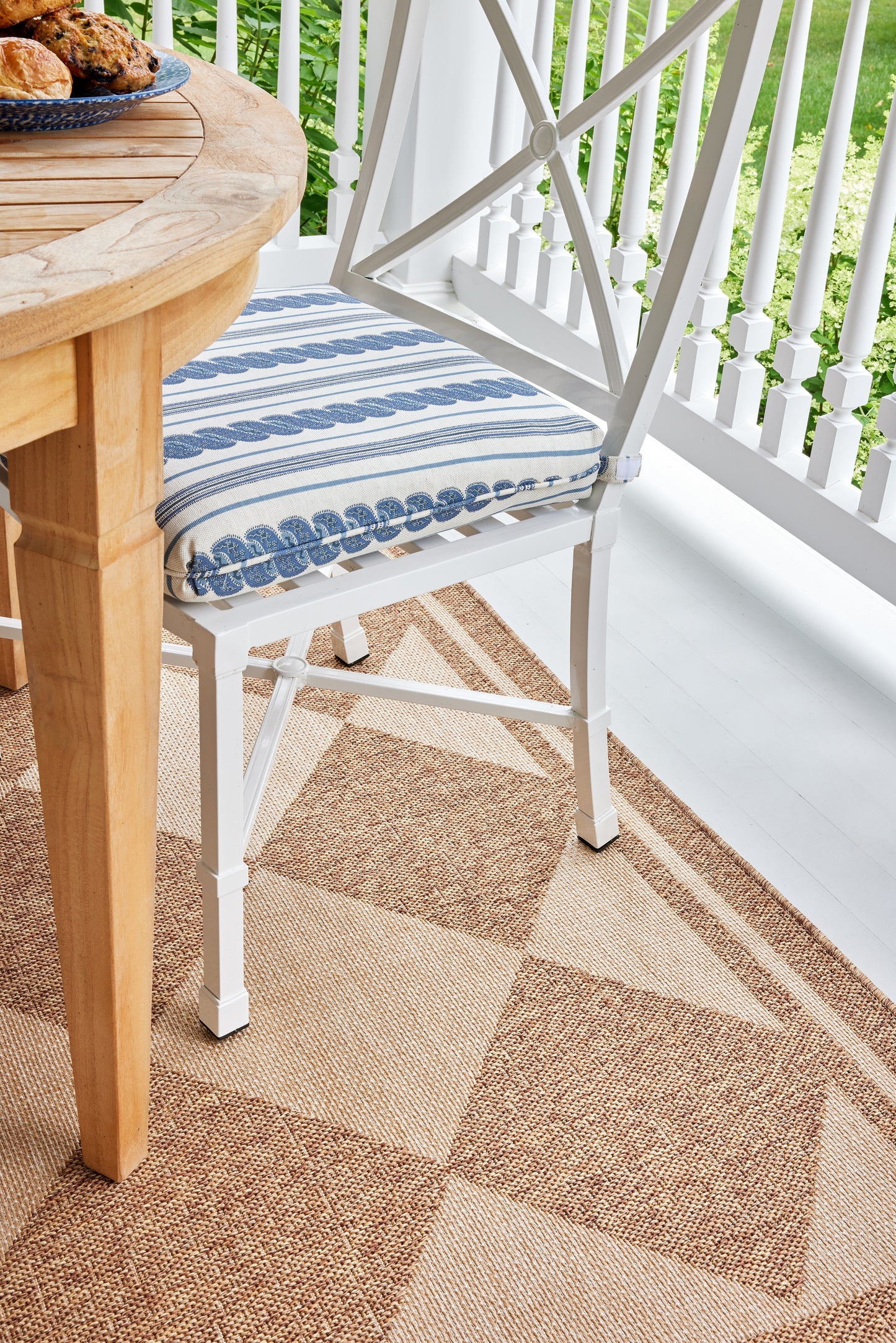 White wrought-iron chair with a blue striped cushion beside a round wooden table on a beige outdoor porch rug.