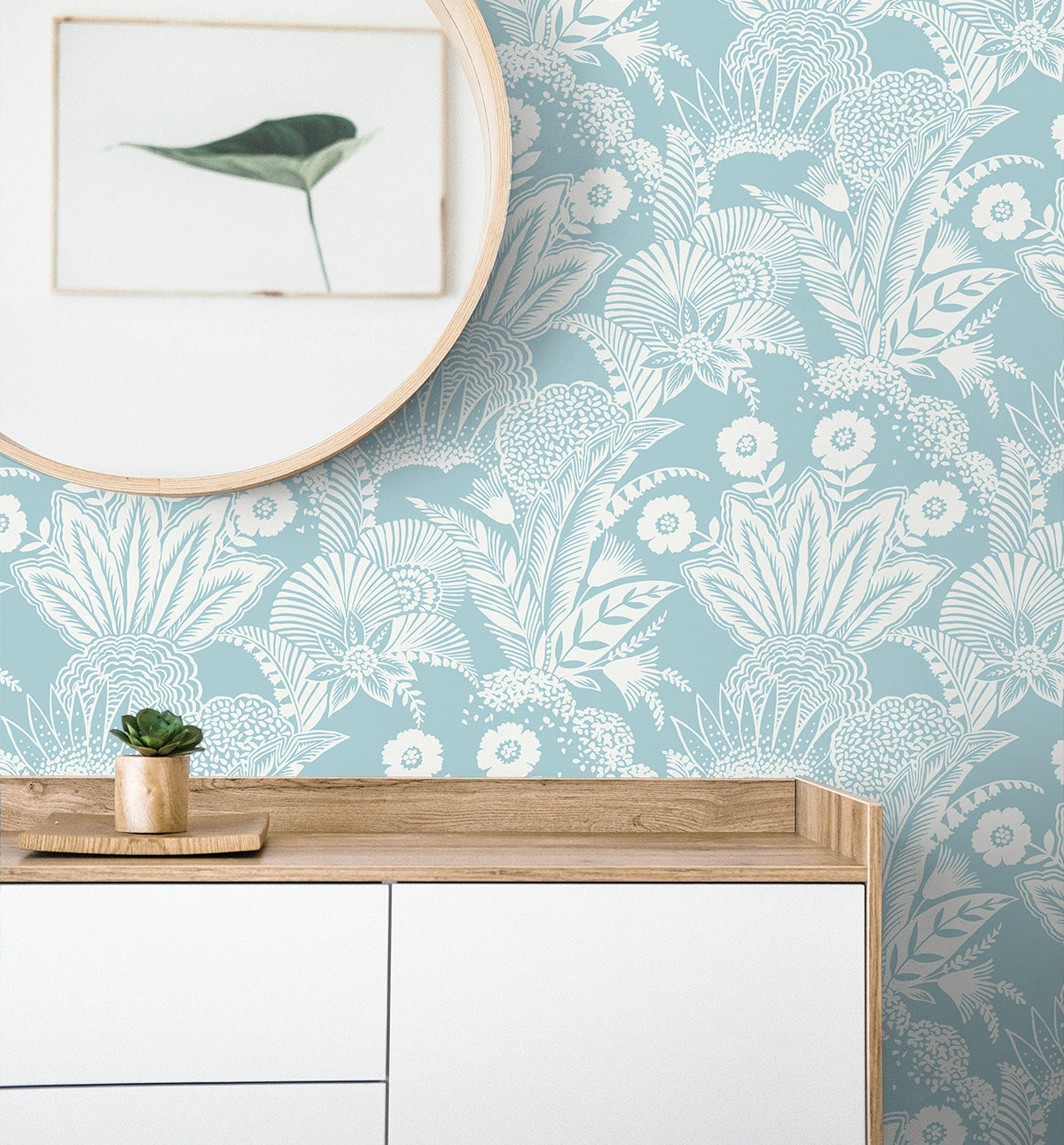 Round wooden-framed mirror over a light wood and white cabinet with a small succulent, blue botanical wallpaper backdrop.