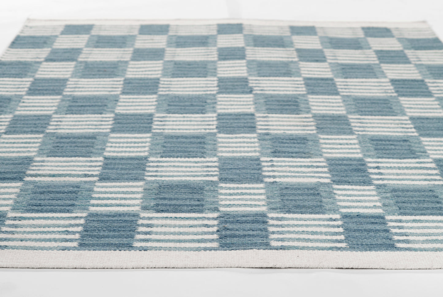 Blue and cream checkerboard woven rug with striped squares; rectangular, contemporary Scandinavian-style decor.