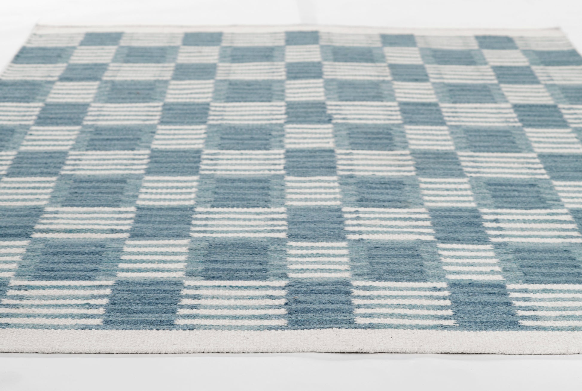 Blue and cream checkerboard woven rug with striped squares; rectangular, contemporary Scandinavian-style decor.