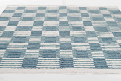 Blue and cream checkerboard woven rug with striped squares; rectangular, contemporary Scandinavian-style decor.
