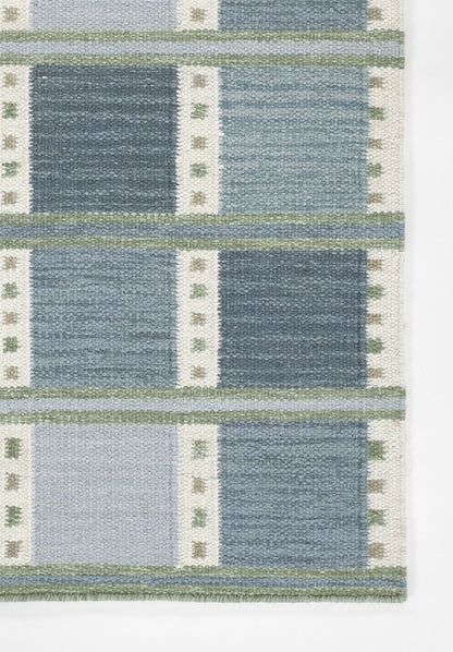 Scandinavian blue woven rug with geometric blocks in navy, light and mid blues, white stripes and green accents.