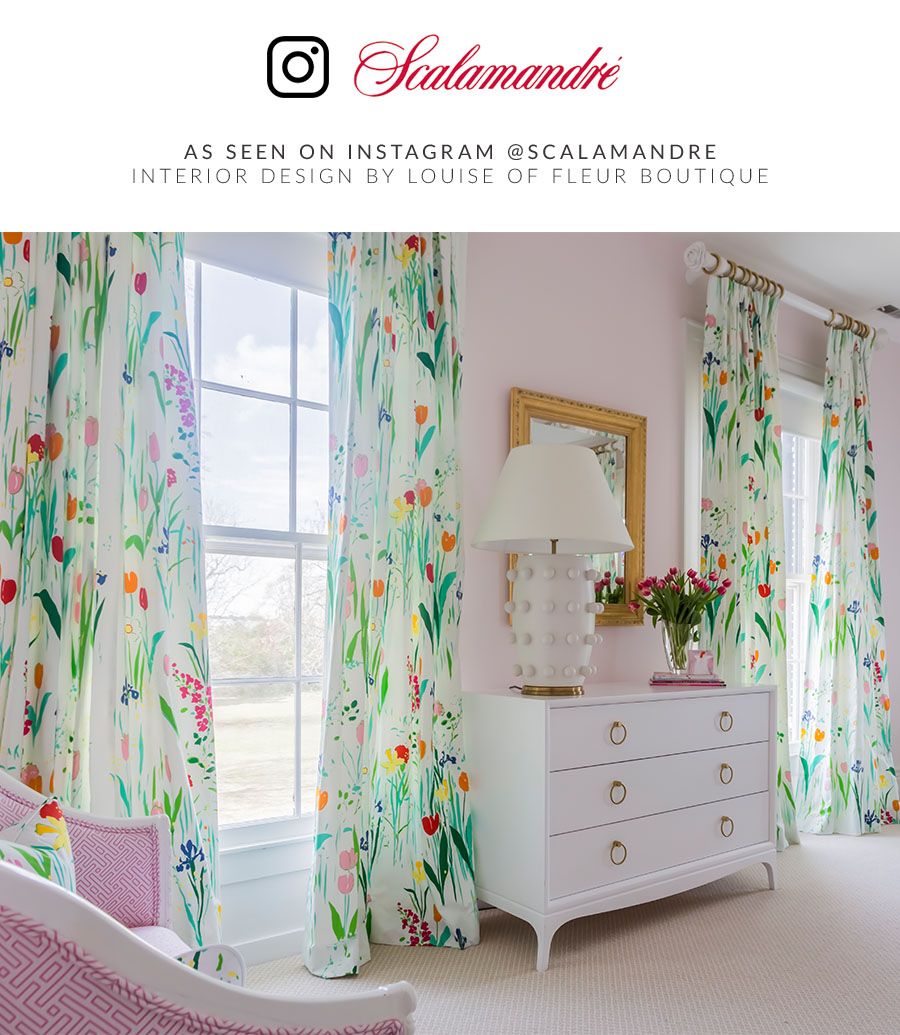 Botanical aqua curtains with multicolored flowers frame a pink bedroom, white curved dresser with gold pulls, and a beaded white lamp.