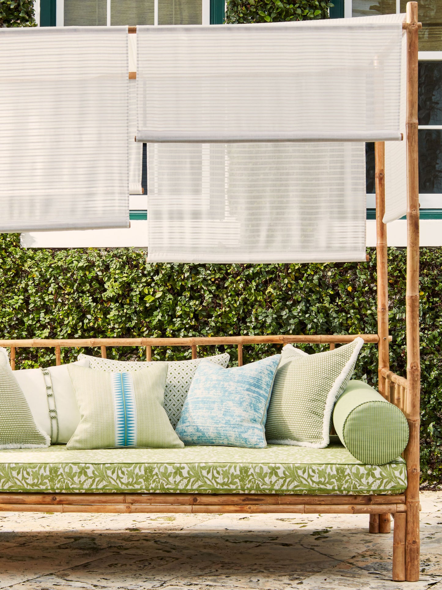 Wooden outdoor daybed with green botanical cushions, blue and cream pillows, and white shade panels on a bamboo frame.