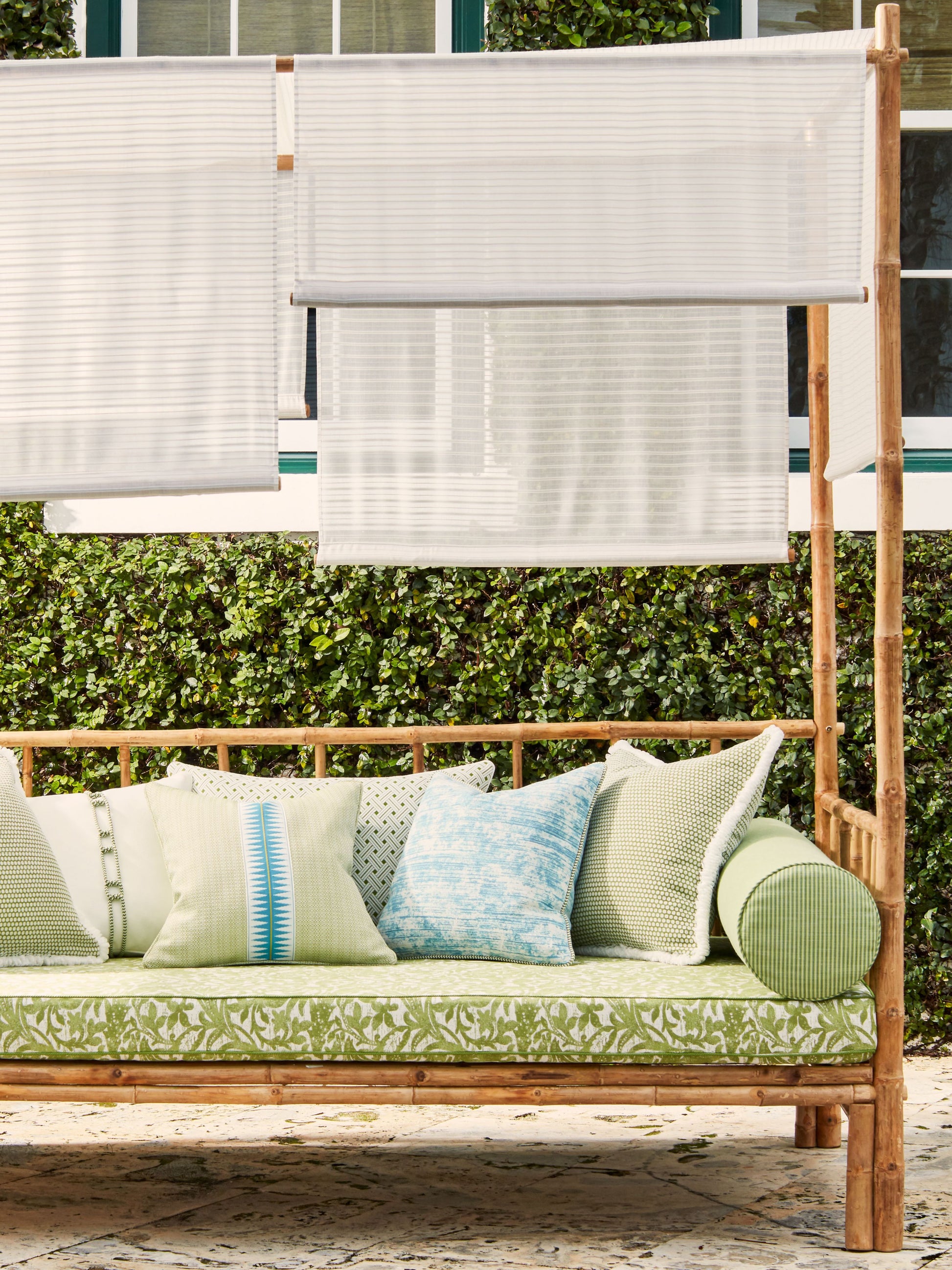 Wooden outdoor daybed with green botanical cushions, blue and cream pillows, and white shade panels on a bamboo frame.