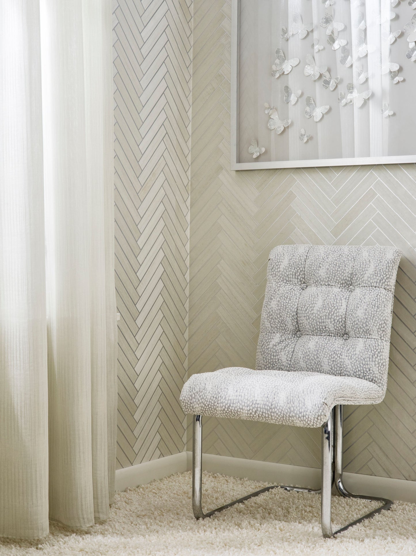 Light gray tufted chair with chrome cantilever legs on a cream shag rug; herringbone wallpaper, sheer curtains.