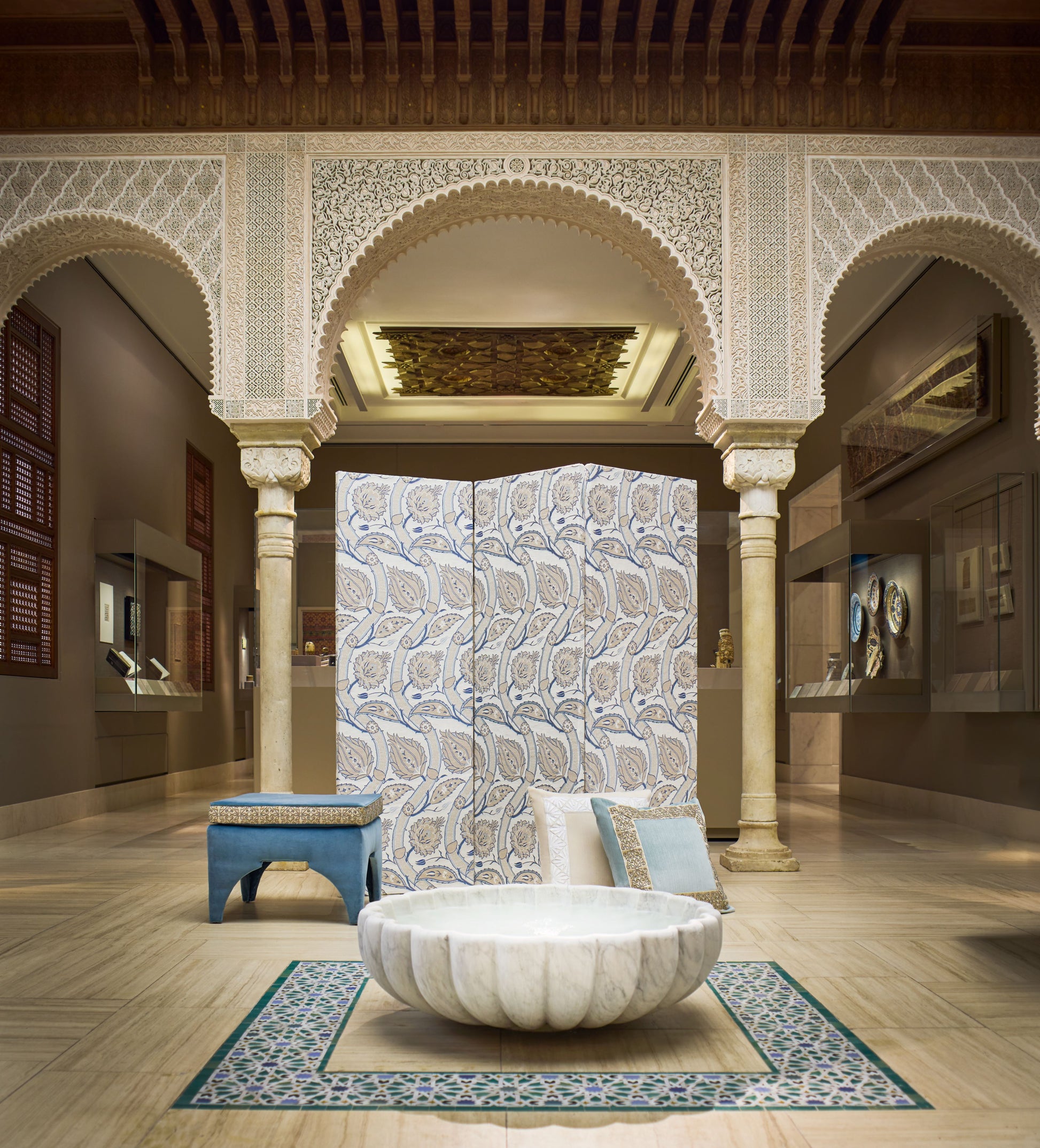 Moroccan-inspired gallery with carved arches, marble columns, blue patterned screen, blue stool, and a round stone bowl centerpiece.