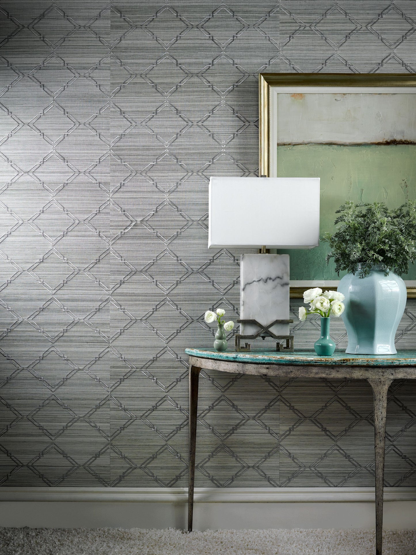 Side-table vignette with lamp on marble-base, turquoise vase, white flowers, and framed art on grey lattice wallpaper.