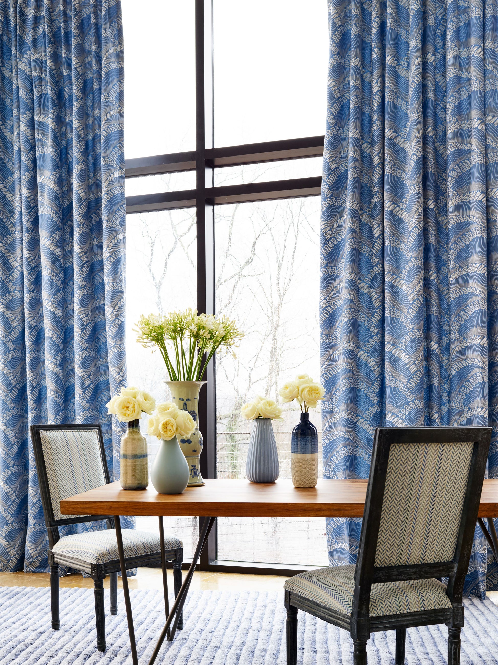 Blue patterned embroidery fabric on display in a room setting, featuring a wooden dining table, white roses in ceramic vases, and two upholstered chairs.