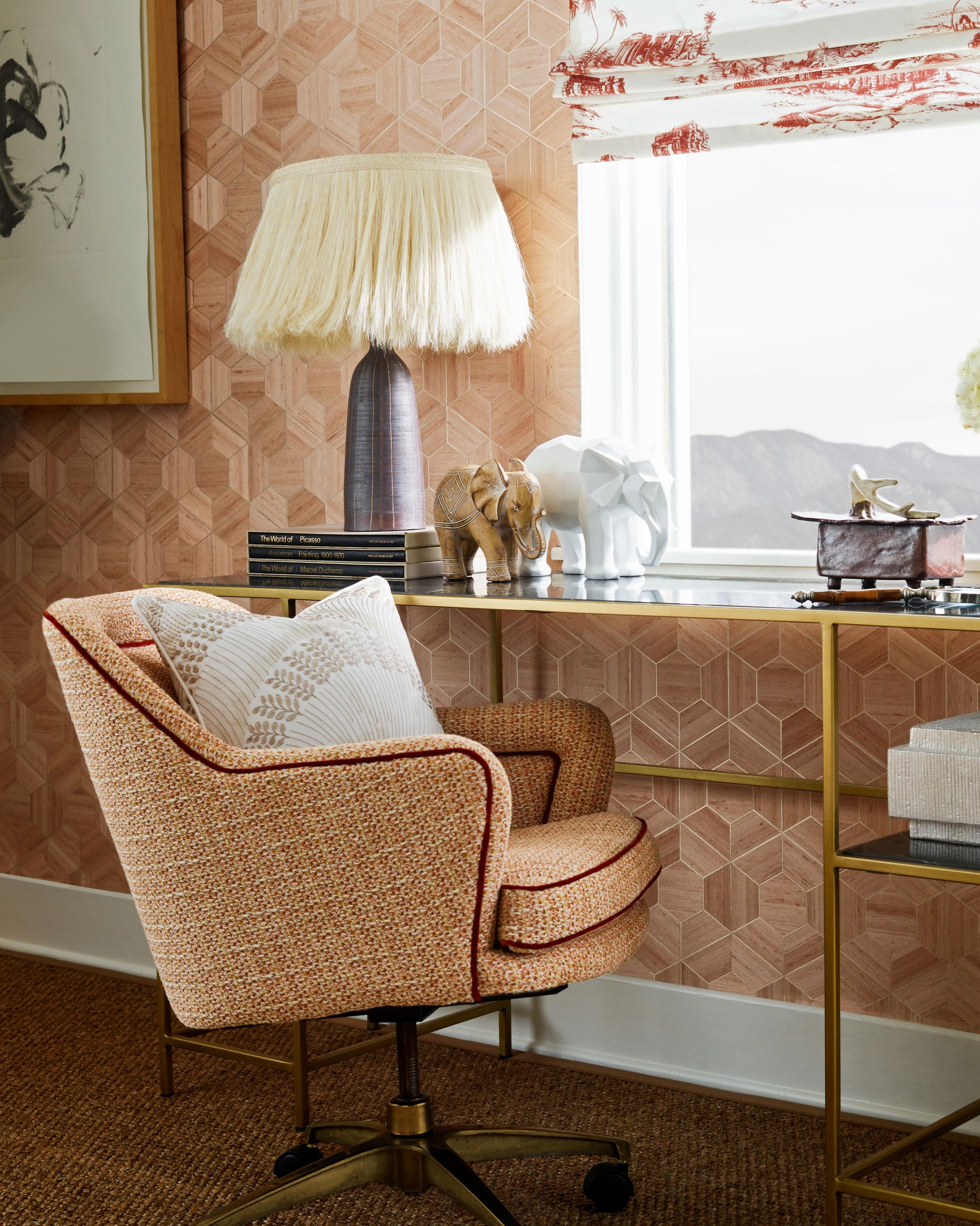 Peach boucle swivel chair, beige fringe lampshade on gold desk; pink geometric wallpaper, elephant decor, window view.