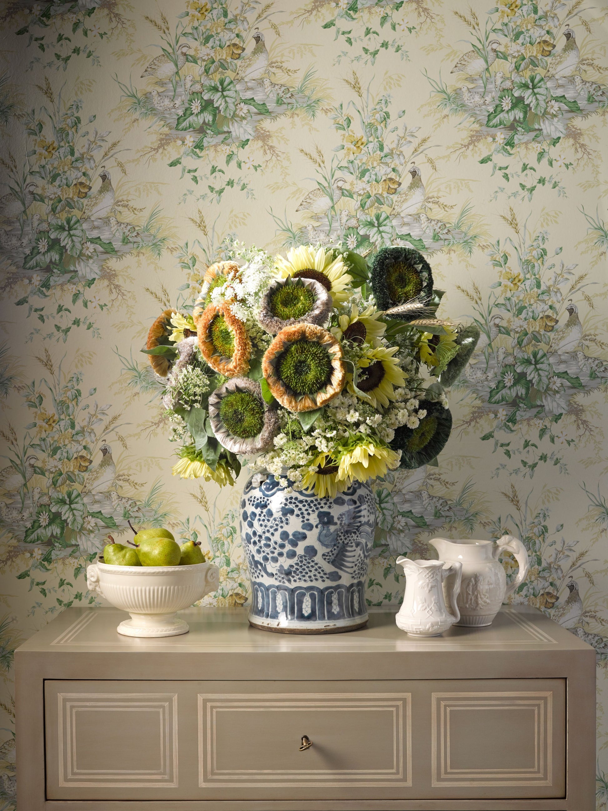 Sunflower bouquet in blue-and-white ceramic vase atop a beige dresser with floral wallpaper; bowl of apples nearby.
