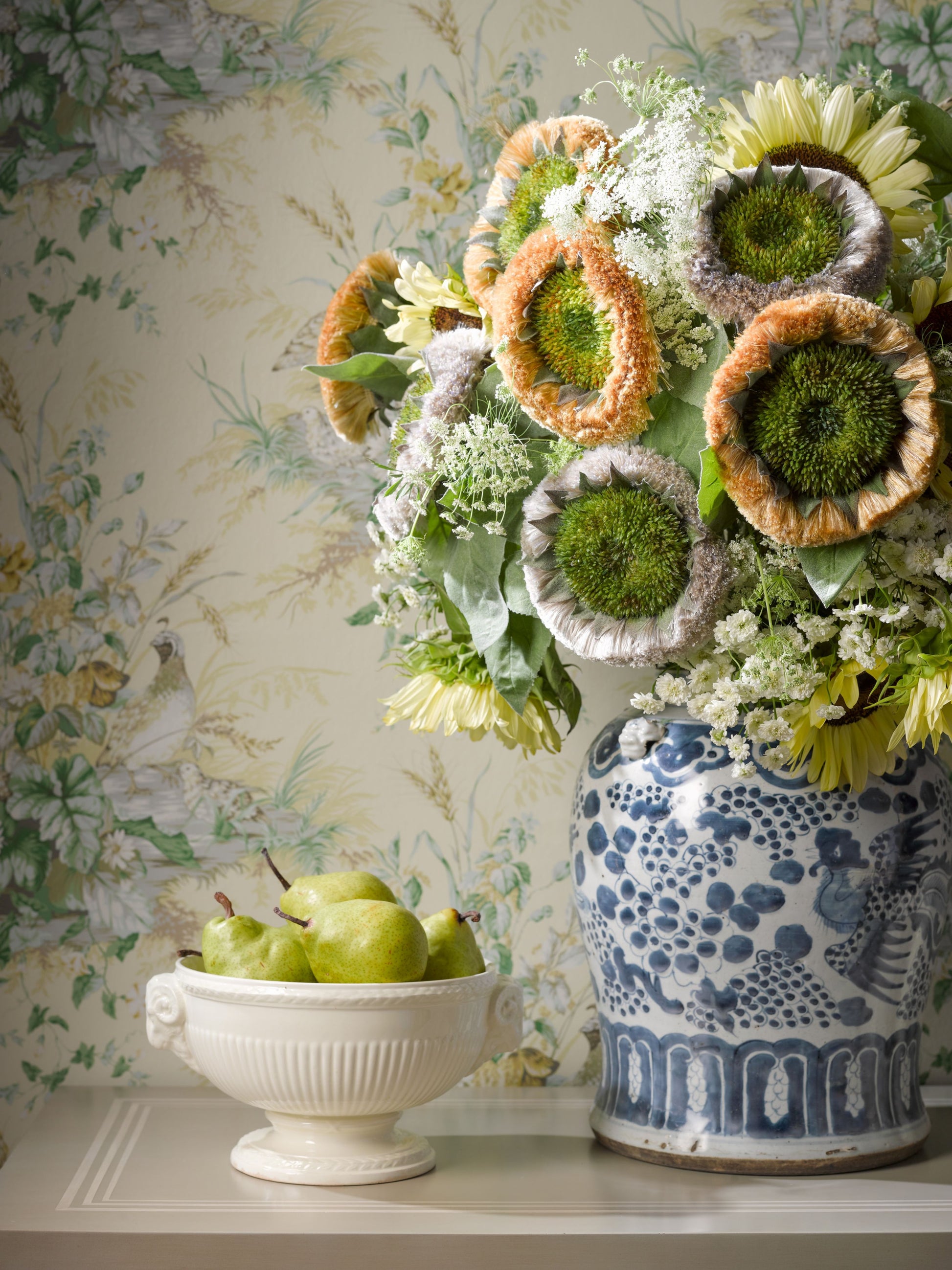Blue and white ceramic vase with sunflower bouquet and dried blooms on a table; white pedestal bowl of green apples nearby.